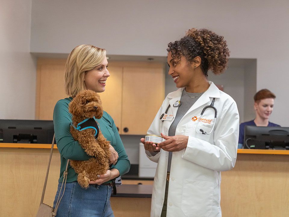 una mujer sonriente que sostiene a un perro negro y habla con una médica veterinaria que también sonríe