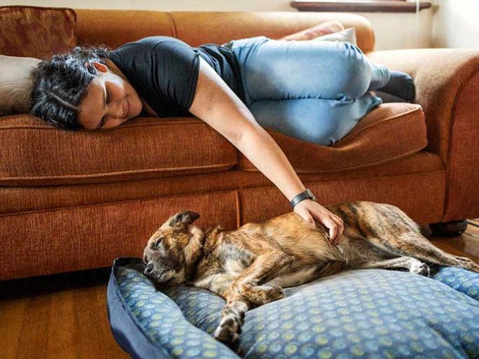 A woman lying on a sofa reaching down and petting a dog lying in a dog bed