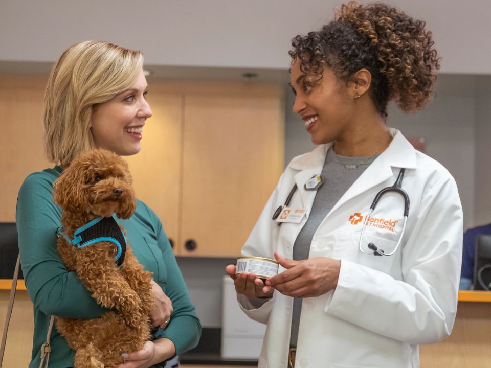 una mujer sonriente que sostiene a un perro marrón y conversa con una médica veterinaria que también sonríe